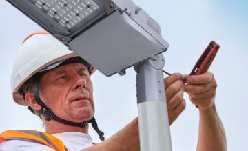 travailleur de la construction portant un casque blanc et un gilet de sécurité orange installe ou répare un lampadaire à l’aide d’un tournevis